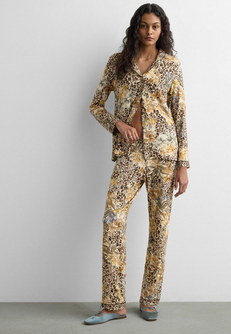 Femme debout contre un mur uni, portant un ensemble de pyjama assorti imprimé floral et léopard, avec des chaussures plates bleu clair.