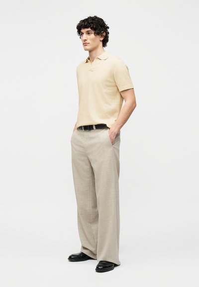 Άνδρας με σγουρά σκούρα μαλλιά, που φοράει ανοιχτό μπεζ κοντομάνικη πόλο, φαρδιά μπεζ παντελόνια, μαύρη ζώνη και μαύρα παπούτσια, στέκεται με τα χέρια στις τσέπες.