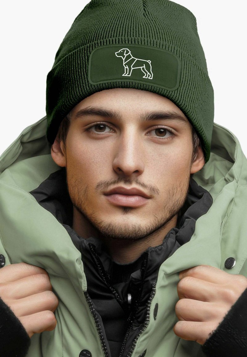 Olive green beanie with a textured ribbed design, featuring a contrasting black patch with a white dog silhouette. Worn with a green jacket.