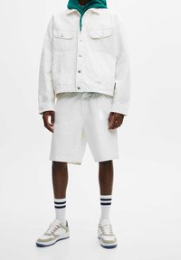 Vit jeansjacka med två framfickor, matchad med vita shorts. Grå och vita sneakers med blå detaljer och randiga träningsstrumpor.