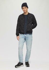 Zwarte gewatteerde bomberjack, marineblauwe trui, lichtblauwe recht gesneden jeans en zwarte sneakers. Model draagt een geribde zwarte beanie.