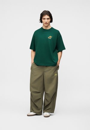 Jauns cilvēks stāv ar rokām kabatās, nēsājot tumši zaļu pārmēru t-kreklu un platus olīvu zaļas krāsas bikses ar baltām sporta kurpēm.