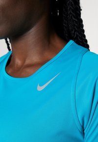 Azurově modré sportovní tričko vyrobené z hladkého, lehkého materiálu. Obsahuje kulatý výstřih a stříbrné logo Nike ve tvaru swoosh na hrudi.