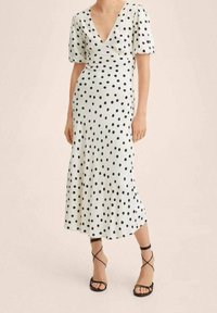 Robe à pois blanche avec des pois noirs, présentant un décolleté en V profond et de courtes manches bouffantes. Fabriquée en tissu lisse avec un ourlet évasé.