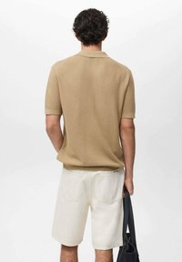 Beige ribbed polo shirt with short sleeves, paired with white denim shorts. The subject holds a black tote bag on the left.