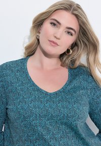 Turkosmönstrad topp med V-ringning, med en texturerad design och långt ärm. Modellen bär guld hoopsörhängen.