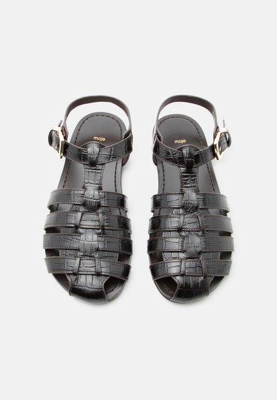 Μαύρα πλεκτά δερμάτινα σανδάλια με λουριά αστραγάλου και χρυσά αγκράφα, επίπεδη σόλα και το όνομα της μάρκας "maje" στην εσωτερική σόλα.