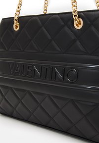 Sac à main en cuir matelassé noir avec une bande de logo en relief, des accessoires en or et des poignées à double chaîne. Surface texturée avec des motifs diagonaux.