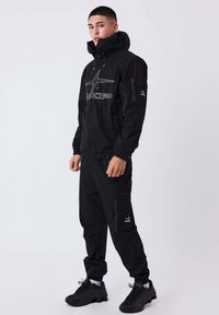 Giovane uomo in giacca con cappuccio nera e pantaloni cargo con logo, in piedi e che guarda leggermente di lato su uno sfondo bianco.