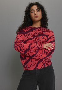 Rot und dunkel lila gemusterter Strickpullover mit langen Ärmeln und geripptem Saum, der ein gewboldes abstraktes Design aufweist. Schwarze Hosen werden getragen.