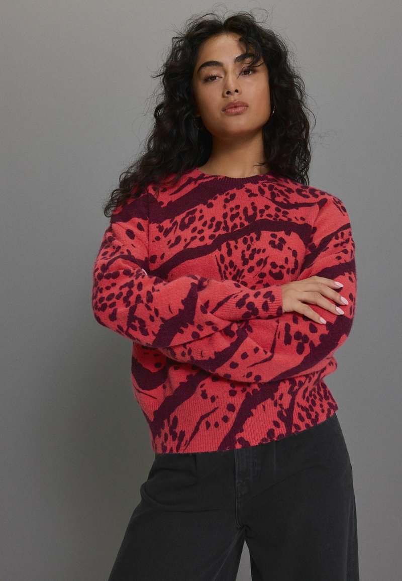 Rot und dunkel lila gemusterter Strickpullover mit langen Ärmeln und geripptem Saum, der ein gewboldes abstraktes Design aufweist. Schwarze Hosen werden getragen.