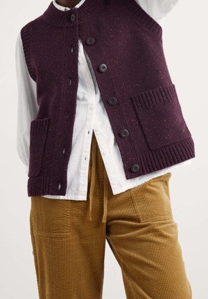 Chaleco de punto morado oscuro abotonado con bolsillos delanteros sobre una camisa blanca, combinado con pantalones de pana marrones.
