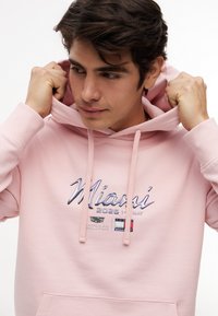 Giovane che sistema il cappuccio di una felpa rosa con la scritta "Miami 2026 1 Maggio" e piccoli loghi stampati sul petto.