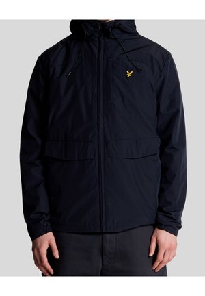 Homme portant une veste noire zippée avec capuche et logo d'oiseau jaune sur la poitrine gauche, debout devant un fond gris uni.