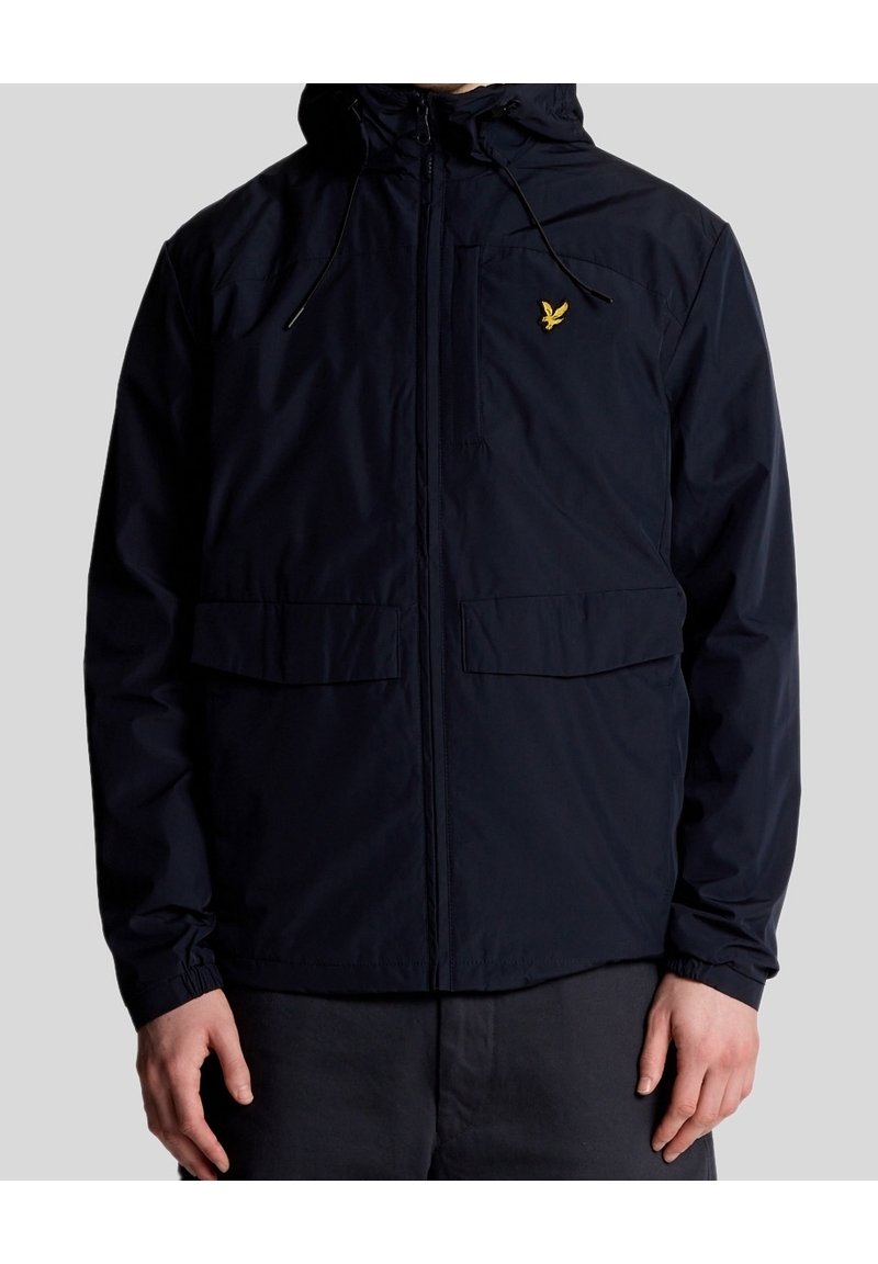 Homme portant une veste noire zippée avec capuche et logo d'oiseau jaune sur la poitrine gauche, debout devant un fond gris uni.