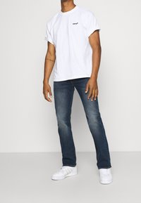 Hombre con una camiseta blanca de Levi's, jeans azules y zapatillas blancas, de pie frente a un fondo claro.