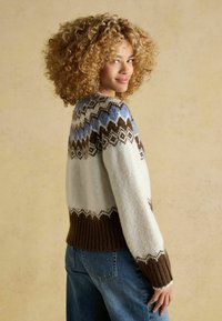 Kobieta z kręconymi włosami, ubrana w wzorzysty wełniany sweter w niebieskch, brązowych i białych kolorach, nosząca niebieskie dżinsy, stojąca na beżowym tle.