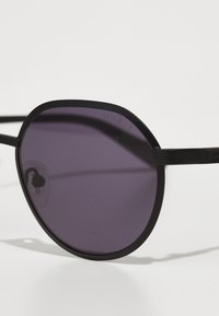 Ochelari de soare rotunzi, negri, cu lentile polarizate de culoare purpurie, având un cadru subțire și elegant, și numele brandului "Guess" discret embosat pe lateral.