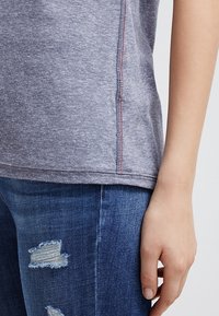 Højre hånd hviler ved siden af slidte blå jeans med revnede lapper, iført en lysegrå skjorte med synlige røde syninger langs sidesømmen.