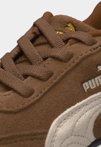 Καφέ σουέντ αθλητικό παπούτσι με καφέ υφασμένες κορδόνια, λευκό λογότυπο Puma στο πλάι και ορατή χρυσή λεπτομέρεια στη γλώσσα.
