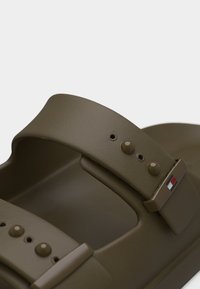 Κοντινό πλάνο σανδαλιού ελιάς με δύο ρυθμιζόμενα λουριά που έχουν στρογγυλά κουμπώματα και μικρό ορθογώνιο λογότυπο.