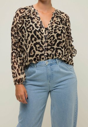 Femme portant un chemisier transparent à motif léopard avec des détails à volants et un jean taille haute bleu clair, debout devant un fond uni.