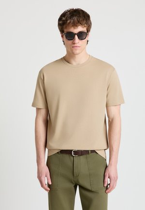Giovane uomo con occhiali da sole neri, t-shirt beige a maniche corte, pantaloni verde oliva e cintura marrone scuro, in piedi davanti a uno sfondo chiaro e uniforme.