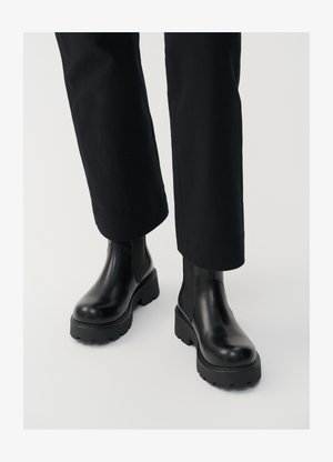 Bottines en cuir noir avec une semelle épaisse, des panneaux latéraux élastiques et une finition lisse, accompagnées de pantalons noirs amples.