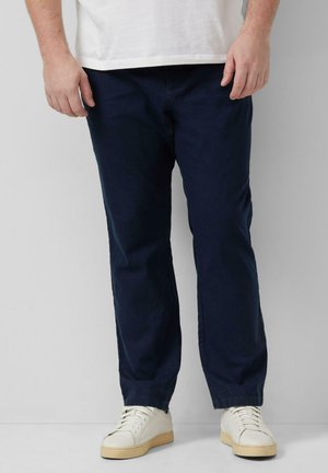 Mann trägt marineblaue Hose und weiße Sneaker, steht vor einem hellgrauen Hintergrund.