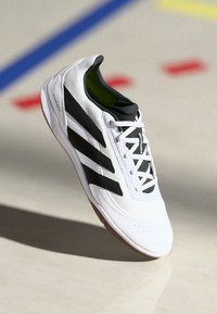 Scarpa sportiva bianca da interno con accenti neri e tre strisce nere.Parte superiore in materiale sintetico testurizzato, suola in gomma e rivestimento verde.