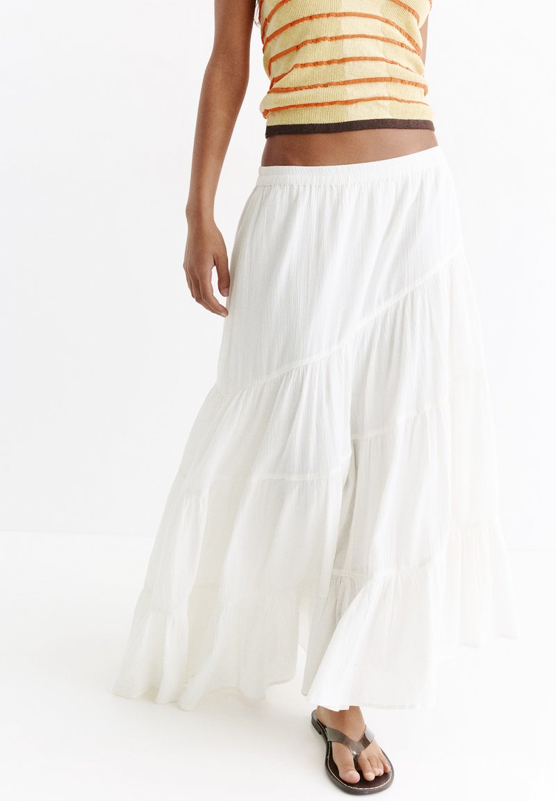 Femme portant une jupe longue blanche à volants, un haut sans manches rayé orange et des sandales noires à brides sur fond blanc.