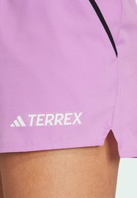 Pantalones cortos deportivos morados hechos de una tela suave, con una abertura lateral y acentos negros. El logo blanco "TERREX" está exhibido de manera prominente.