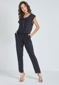 Mono negro hecho de tela ligera, con parte superior de mangas cortas y botones, cinturilla elástica y piernas ajustadas. Sandalias plateadas.