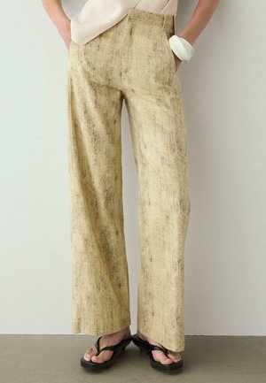 Beżowe spodnie o szerokich nogawkach z teksturą, noszone z czarnymi sandałami na obcasie, jasną bluzką i białą bransoletką na prawej ręce.