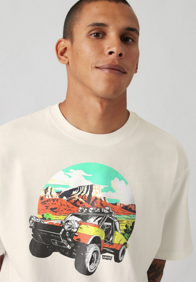 Camiseta color crema con un estampado gráfico de un buggy de arena amarillo y naranja con un paisaje montañoso de fondo.