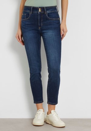 Kobieta w ciemnoniebieskich dopasowanych dżinsach i białych trampkach Guess, stojąca przy gładkiej ścianie.