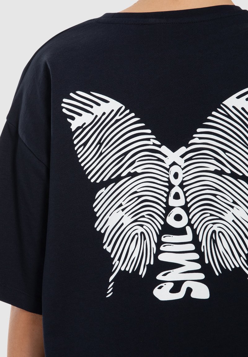 Diseño de mariposa en la parte trasera de una camiseta negra con alas formadas por patrones de huellas dactilares blancas y la palabra "SMILDOOD" en vertical en el centro.