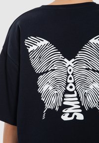 Schwarzes Baumwoll-T-Shirt mit einem großen weißen Schmetterlingsgrafik, die aus Fingerabdrucklinien besteht, wobei das Wort "SMILODOX" in das Design integriert ist.