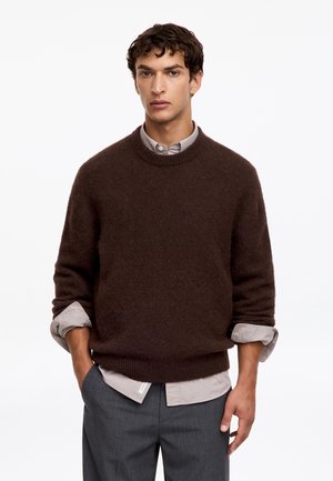 Jeune homme portant un pull marron foncé par-dessus une chemise claire boutonnée avec les manches retroussées et un pantalon gris, debout avec une main dans la poche.