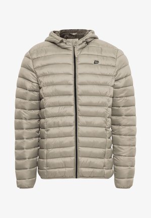 Joven vestido con una chaqueta de plumas beige, camiseta blanca, pantalones negros y zapatillas negras, de pie con las manos relajadas a los lados.