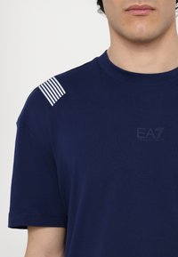 Granatowy t-shirt z bawełny z krótkimi rękawami, z białymi paskami na ramionach oraz subtelnym logo na wysokości klatki piersiowej.