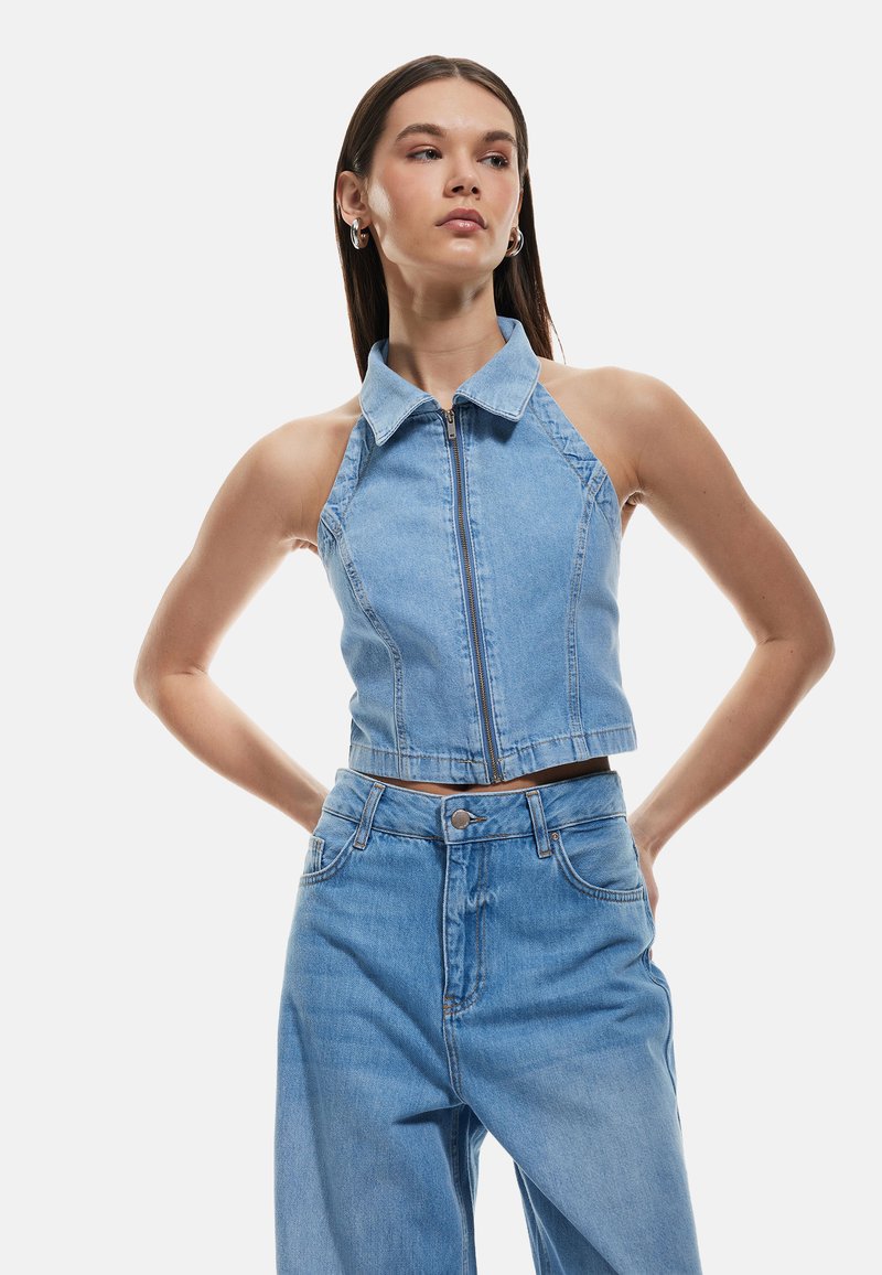 Chaleco de mezclilla de color azul claro, cortado, con cuello y cremallera frontal, que presenta un diseño ajustado y una cintura ajustable, combinado con jeans de cintura alta.