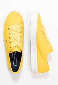 Żółte tenisówki z materiału płóciennego z białymi gumowymi podeszwami i złotymi oczkami. Posiadają sznurowadła w paski oraz logo po boku. Nowoczesny, niski fason.