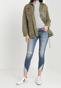 Chaqueta bordada verde oliva con bolsillos, suéter de cuello alto gris, jeans ajustados azul desgastados y zapatillas blancas con detalles en negro y rojo.