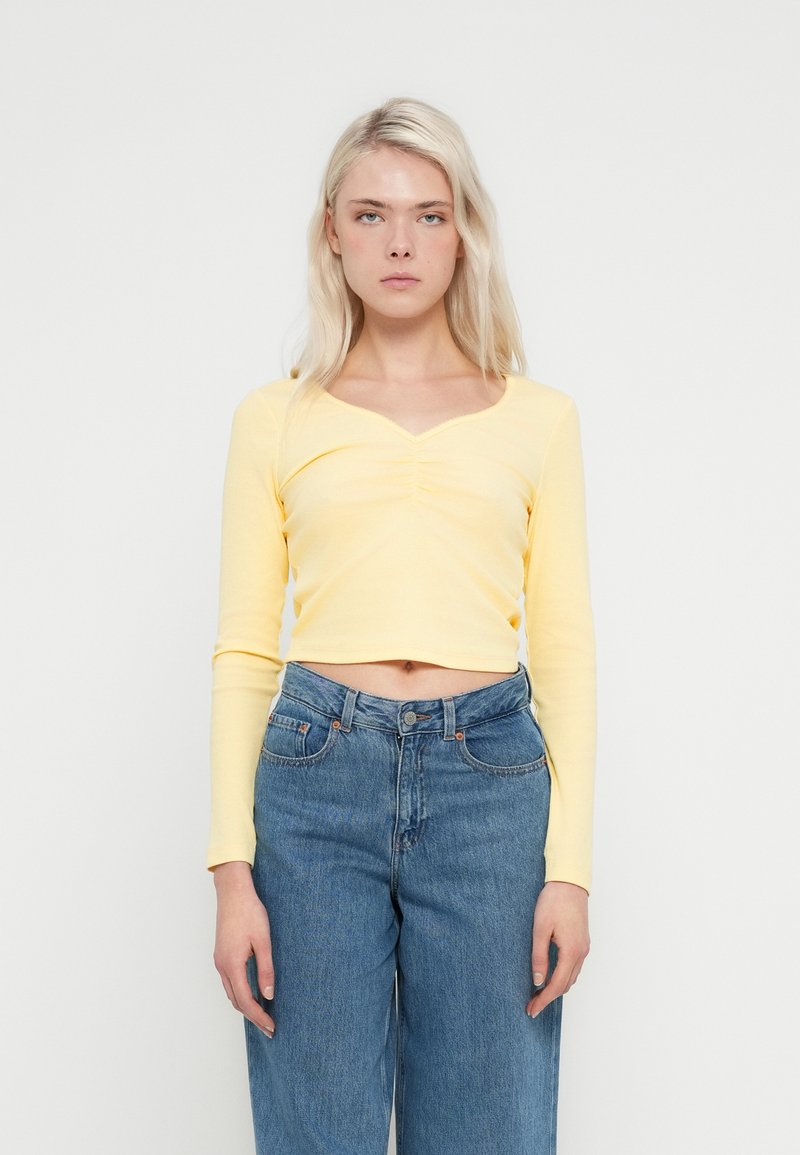 Żółty krótki top z długim rękawem o prążkowanej fakturze i dekoltem w kształcie litery V, zestawiony z wysokim stanem niebieskich dżinsów.