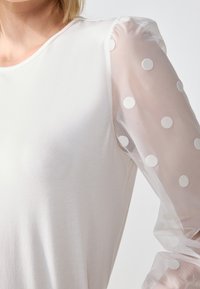 Bílé tričko s měkkým tělem a průhlednými rukávy s bílými puntíky, zvýrazněné mírným nafouknutím na ramenou.
