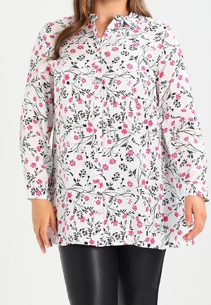 Femme portant un chemisier blanc à manches longues avec un motif floral noir et rose et un pantalon noir, présentée des épaules jusqu’au milieu des cuisses.