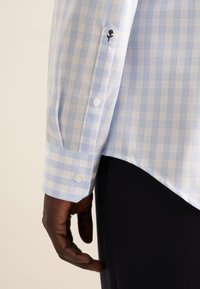Camicia a quadri blu chiaro con accenti bianchi, caratterizzata da un polsino a forma esagonale, chiusura con bottoni e dettaglio ricamato di un fiore sulla manica.