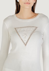 Suéter blanco de punto con un logotipo triangular y embellado de "GUESS" en el centro, acentuado con rhinestones de color claro y un escote redondeado.