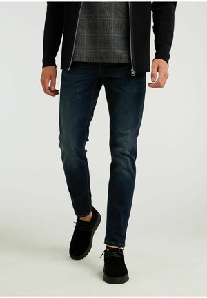 Man wearing dark blue skinny jeans, black shoes, a black zippered jacket, and a gray plaid shirt walking on a white background.